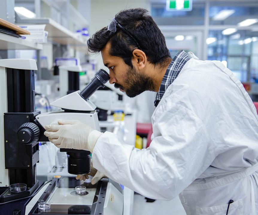 A man wearing a white lab coat and gloves looks into a microscope.